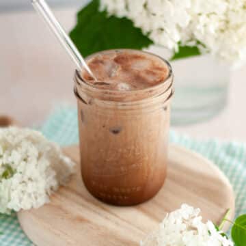 mason jar filled with a frothy iced chocolate shaken espresso with a glass straw. the glass sits on a wooden board and is surrounded by a blue and white checkered linen and hydrangea flowers.