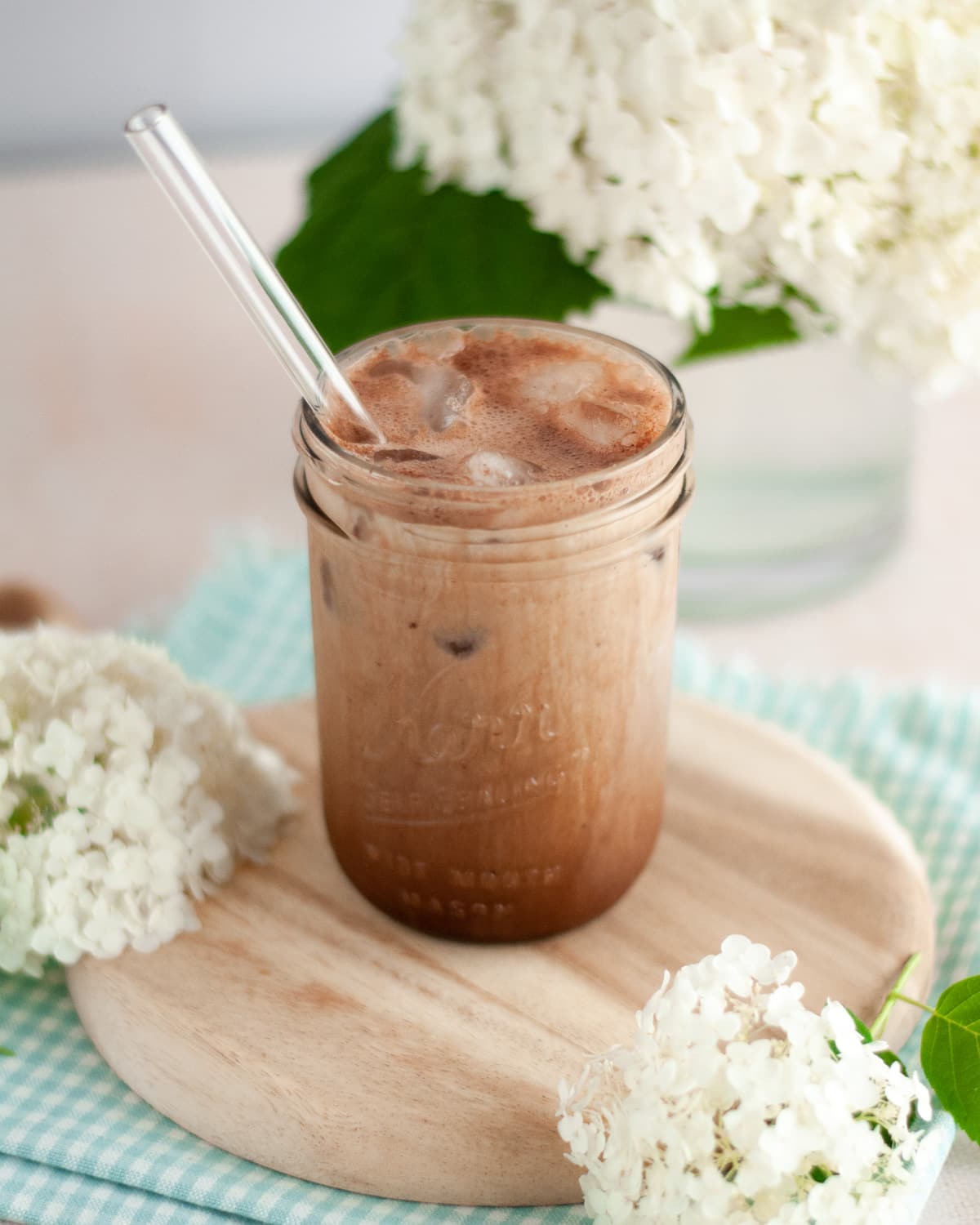mason jar filled with a frothy iced chocolate shaken espresso with a glass straw. the glass sits on a wooden board and is surrounded by a blue and white checkered linen and hydrangea flowers.
