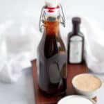 a glass bottle of homemade vanilla syrup sitting on a wooden board, surrounded by ingredients and a white linen.
