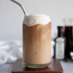 a vanilla sweet cream cold brew coffee served in a tall glass. vanilla sweet cream cold foam sits on top of the iced coffee, with legs of cream dripping down into the coffee. the glass sits on a wooden board, with coffee beans around the scene. a white linen, vanilla extract bottle, and container of cold brew sit in the background.