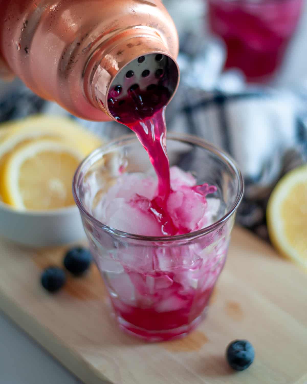 Freshly shaken blueberry vodka lemonade being poured out of a cocktail shaker into a lowball glass filled with ice.