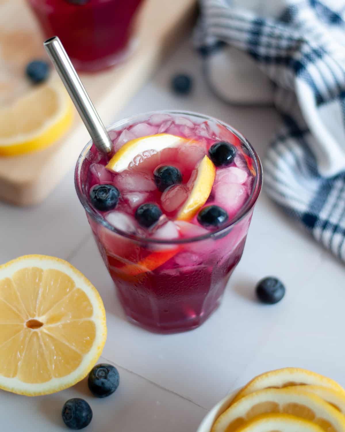Blueberry Vodka Lemonade in a lowball glass, garnished with fresh blueberries and lemon slices. The glass is surrounded by ice, blueberries, lemon wheels, and a blue and white checkered linen.