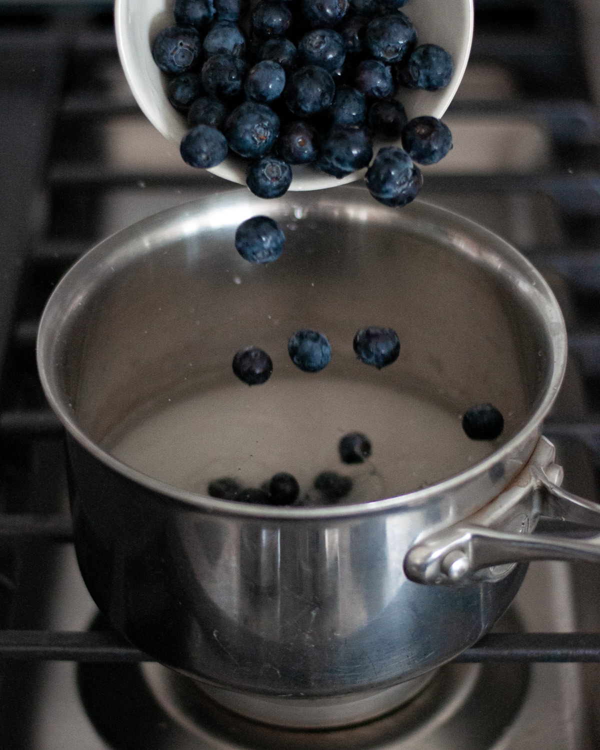 process shot for making blueberry simple syrup. this shot shows fresh blueberries being added to water and granulated sugar being in a medium sized saucepan on the stovetop.