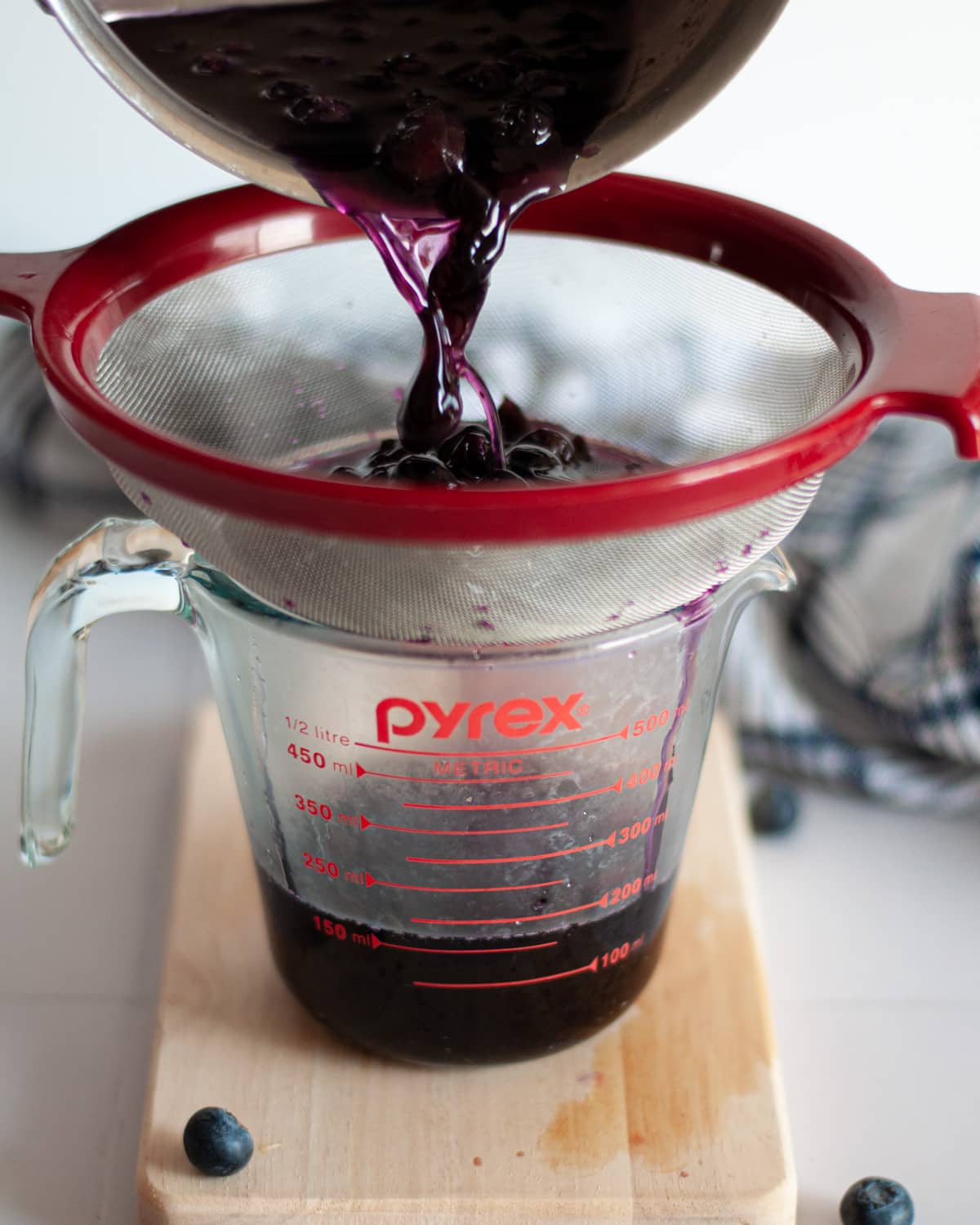 process shot for making blueberry simple syrup. this shot shows the blueberry syrup being strained through a fine mesh sieve to remove the blueberries.