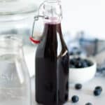 glass container for deep purple blueberry syrup. fresh blueberries are scattered around the scene. a container of sugar and navy and white checkered linen sit in the background, with a glass container of water in the foreground.