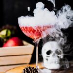 Tabletop view of a smoking Poison Apple Cocktail on a wooden board with a skull and a pinecone decoration on the table beside the glass.