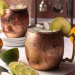 Close-up shot of a Pumpkin Gin Mule cocktail in a copper mug, a slice of lime for the garnish, and additional slices of lime scattered throughout the photo.