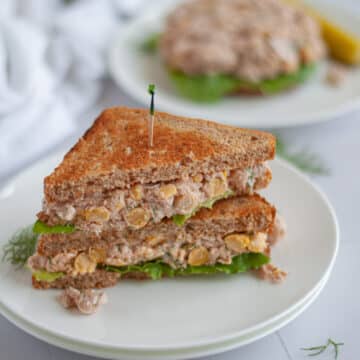 a healthy tuna salad sandwich cut in half and stacked on top of each other on a stack of white plates. an open-faced tuna and chickpea salad sandwich sits in the background.