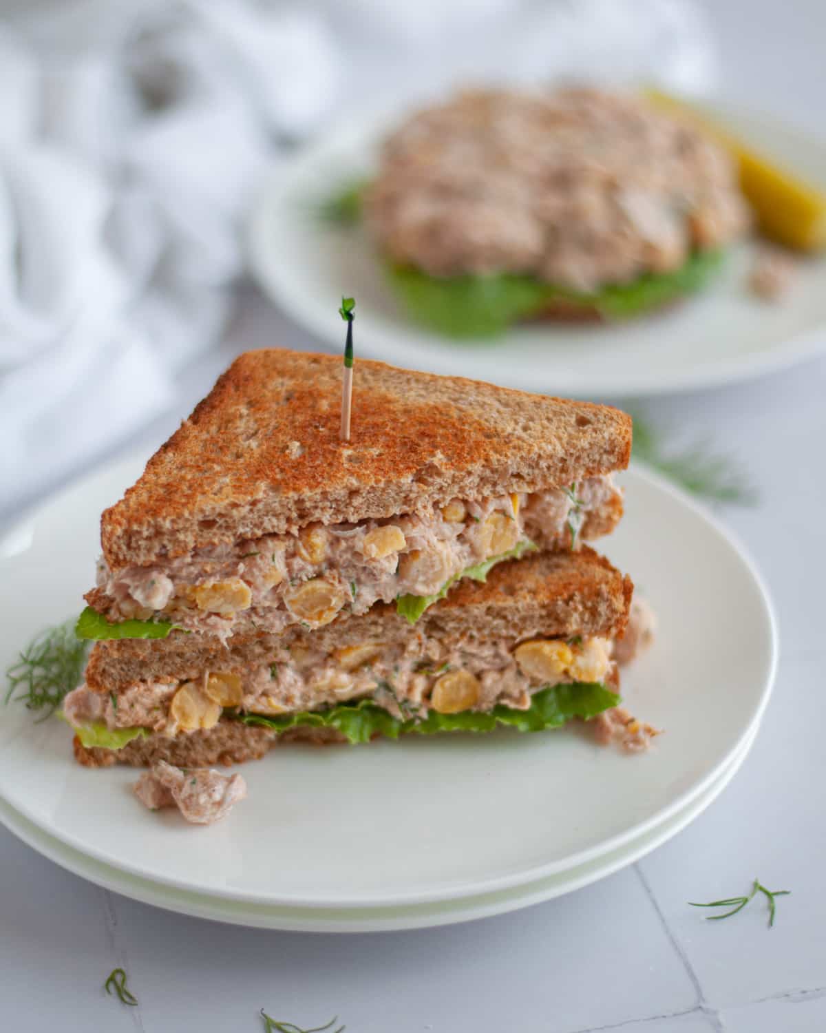 a healthy tuna salad sandwich cut in half and stacked on top of each other on a stack of white plates. an open-faced tuna and chickpea salad sandwich sits in the background.