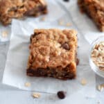 a oatmeal chocolate chip cookie bar sits on top of a piece of parchment paper with rolled oats and chocolate chips scattered around it. additional cookie bars sit throughout the scene.