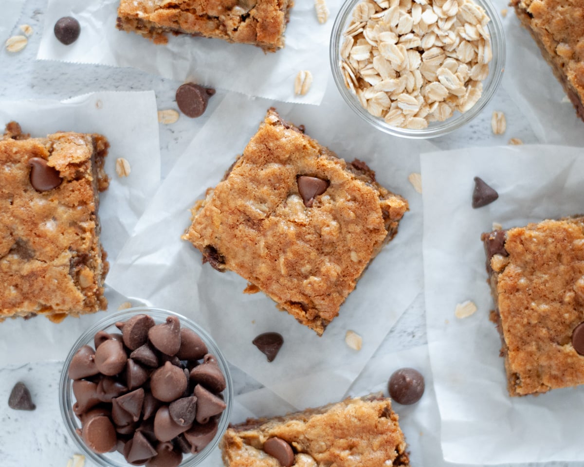 top down view of oatmeal chocolate chip cookie bars that are sitting on squares of parchment paper with rolled oats and chocolate chips scattered around them.