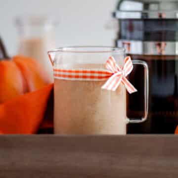 a glass pitcher filled with homemade pumpkin spice creamer. the pitcher has a orange and white checkered ribbon tied to it, and is sitting on a wooden tray with an orange linen, a pumpkin, and a french press filled with hot coffee.
