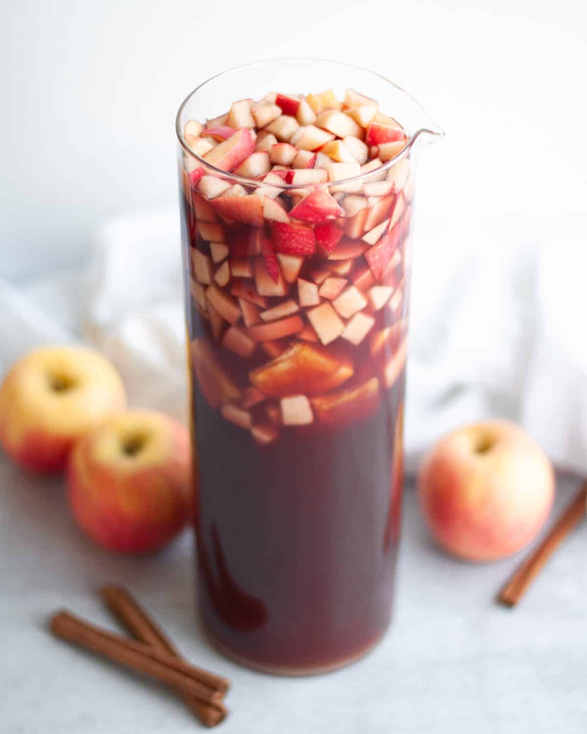 Tall glass pitcher of fall red wine sangria. A white linen, apples, and cinnamon sticks sit around the pitcher.