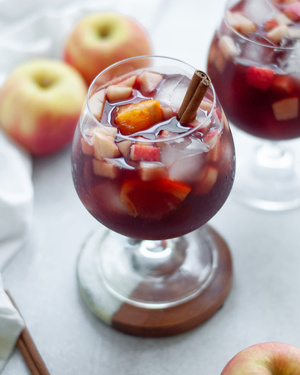 Close up shot of two wine glasses filled with fall red wine sangria and garnished with a cinnamon stick. Apples and a white linen are in the background.