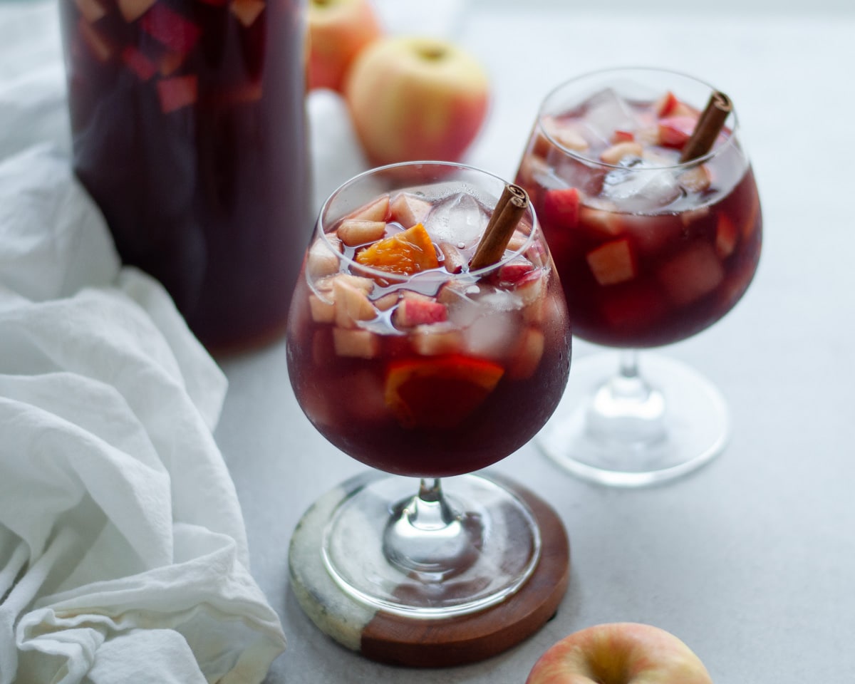 Close up shot of two wine glasses filled with fall red wine sangria and garnished with a cinnamon stick. The pitcher of sangria is in the background, with apples and a white linen.