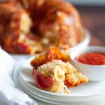 pieces of pepperoni pull apart bread sit on a plate with the bowl of sauce, and the rest of the monkey bread is in the background.