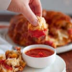 a piece of pizza monkey bread that has just been dipped into pizza sauce. additional pieces of monkey bread sit on a plate with the bowl of sauce, and the rest of the monkey bread is in the background.