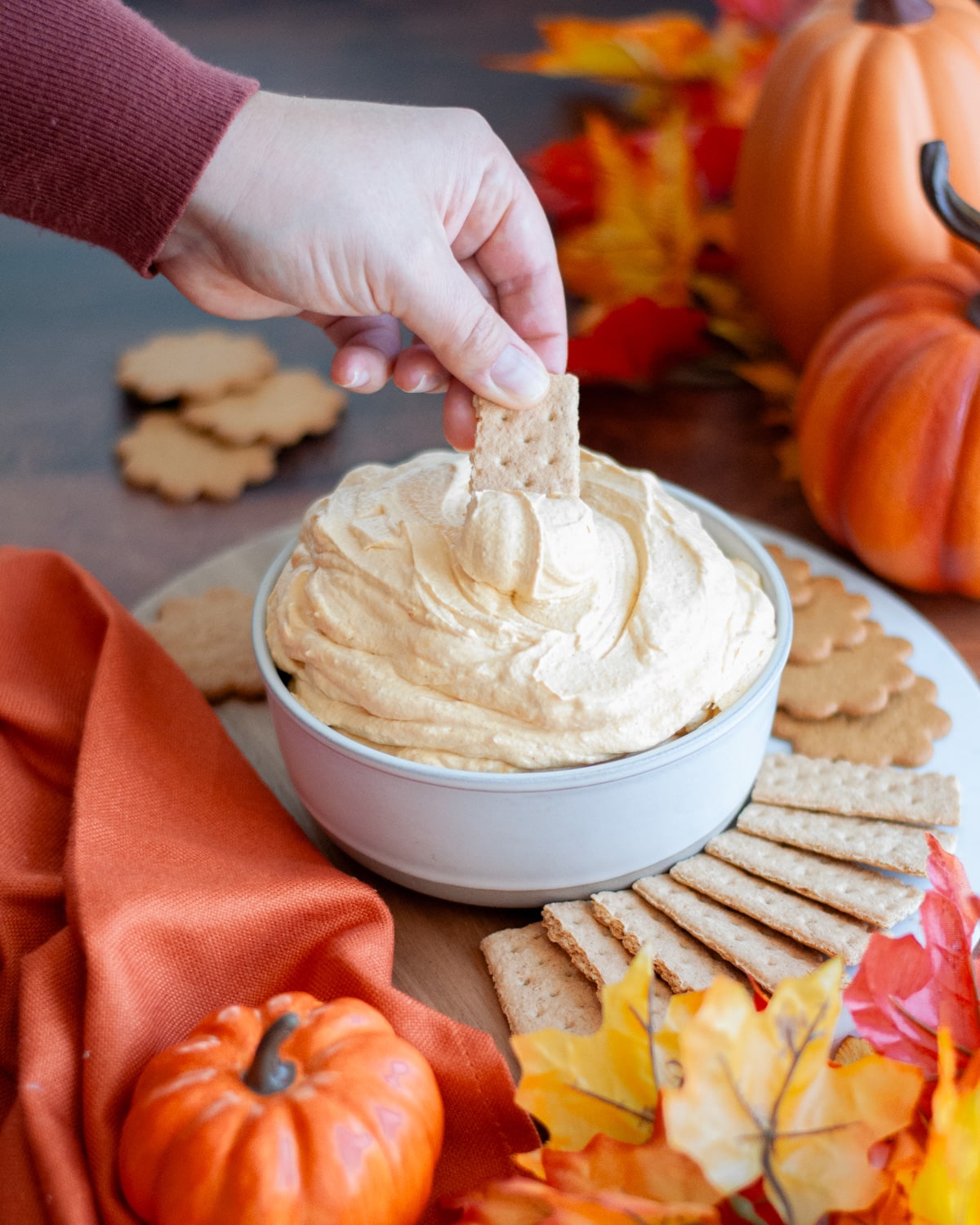 a hand dipping a graham cracker into a serving bowl filled high with pumpkin dip sitting on top of a serving board filled with gingersnaps and graham crackers. the treat is surrounded by an orange linen, pumpkins, and fall leaves.