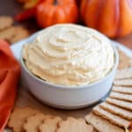 a serving bowl filled high with pumpkin dip sitting on top of a serving board filled with gingersnaps and graham crackers. the treat is surrounded by an orange linen, pumpkins, and fall leaves.