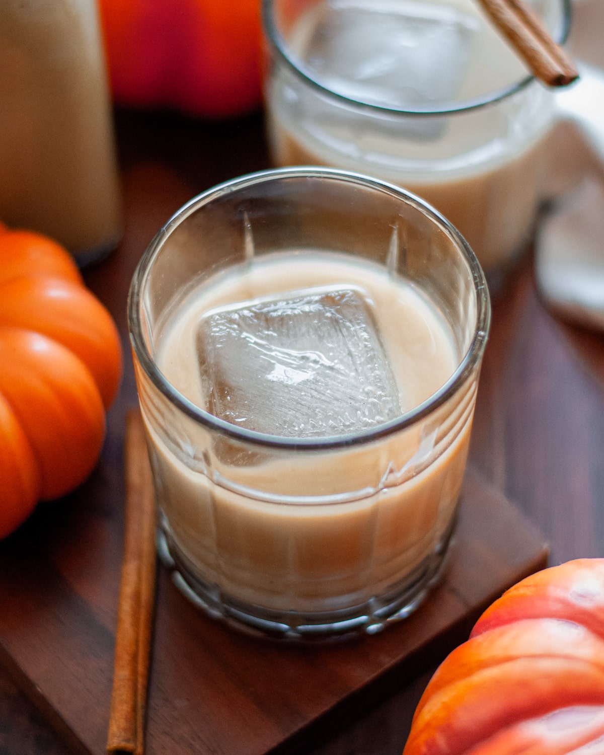an old fashioned glass filled with a pumpkin spice white russian cocktail with a large ice cube and a cinnamon stick. the cocktail sits on a wooden board with pumpkins around it, and another cocktail in the background.