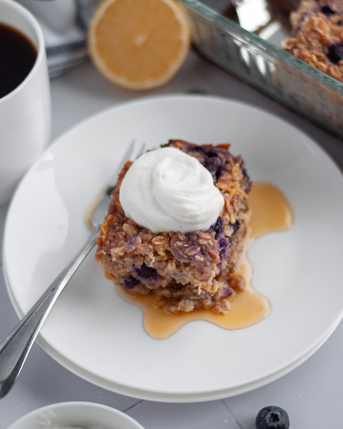 A slice of blueberry lemon baked oatmeal on a white plate with maple syrup and a dollop of vanilla Greek yogurt on top.
