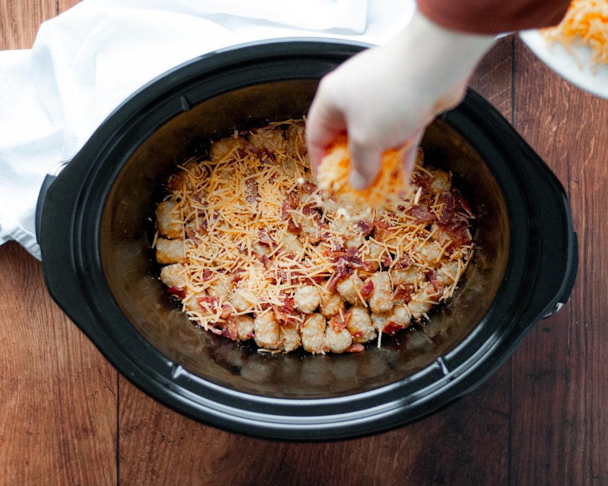 Process shot showing how to make tater tot and chicken casserole in the slow cooker, showing the bottom a crockpot lined with a single layer of tater tots, bacon, and cheese being added.