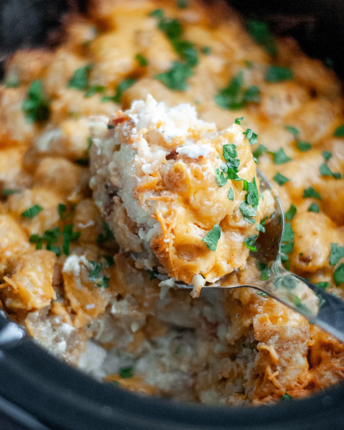 Close up of a serving spoon with a scoop of the ranch bacon chicken and tater tot casserole on it, sitting on top of the rest of the casserole in the crockpot basin.