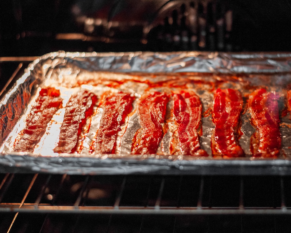Process shot showing bacon cooking on a lined baking sheet on the center rack of the oven.