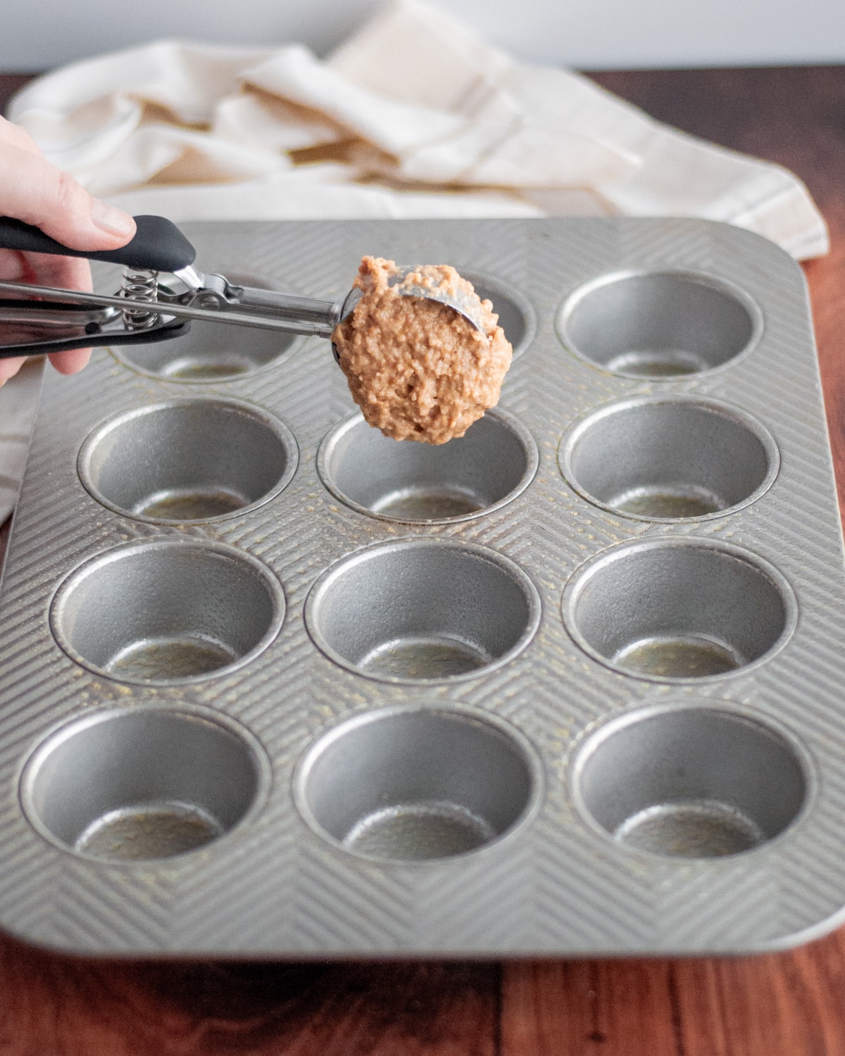 Process shot showing how to make wheat germ muffins; image shows the muffin batter being spooned into a muffin tin with a large cookie scoop.