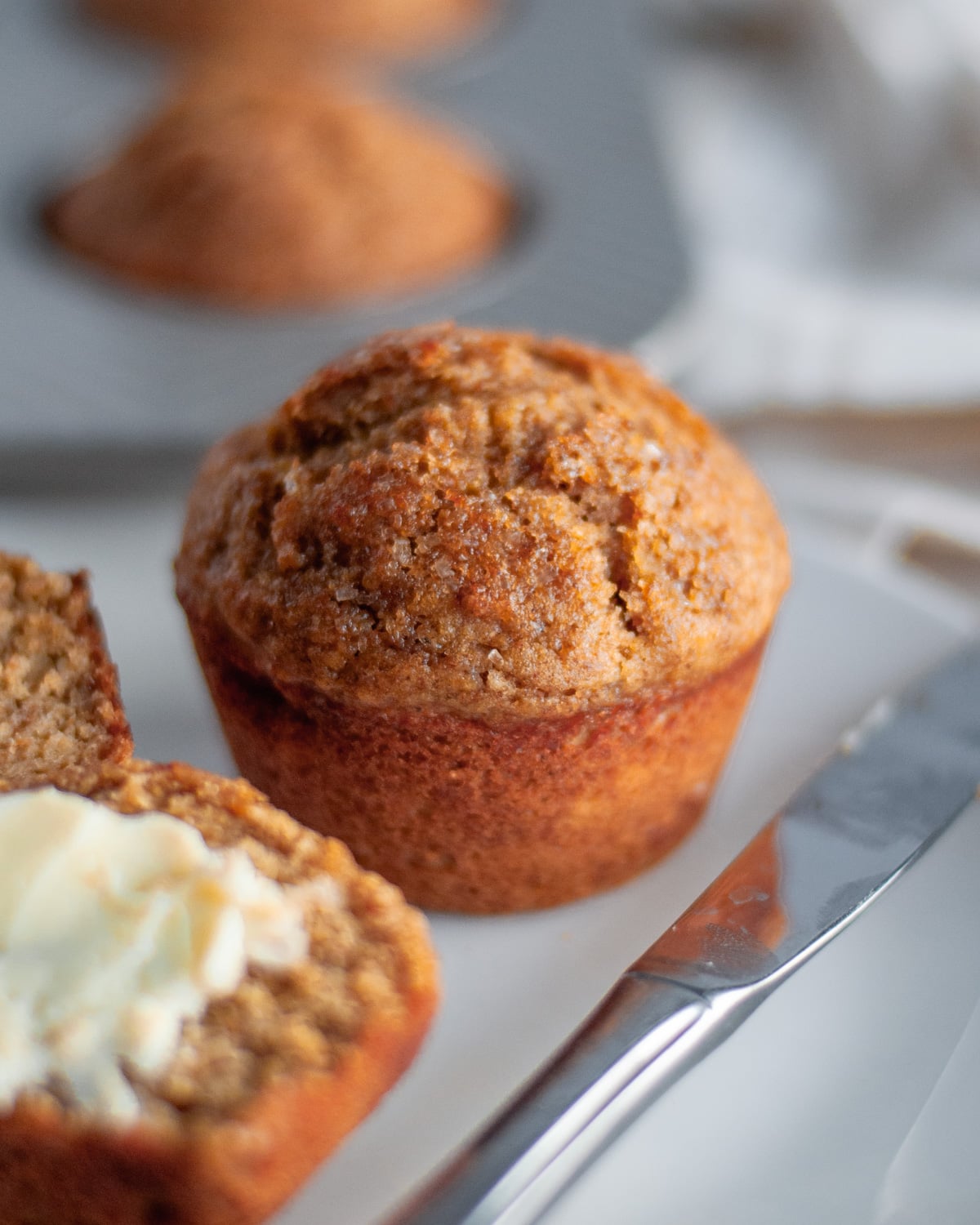 Close up of a full wheat germ muffin on a white serving plate with a buttered muffin in the foreground.