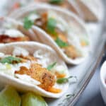 A long tray of crispy fish tacos topped with slaw, cilantro, and salsa.