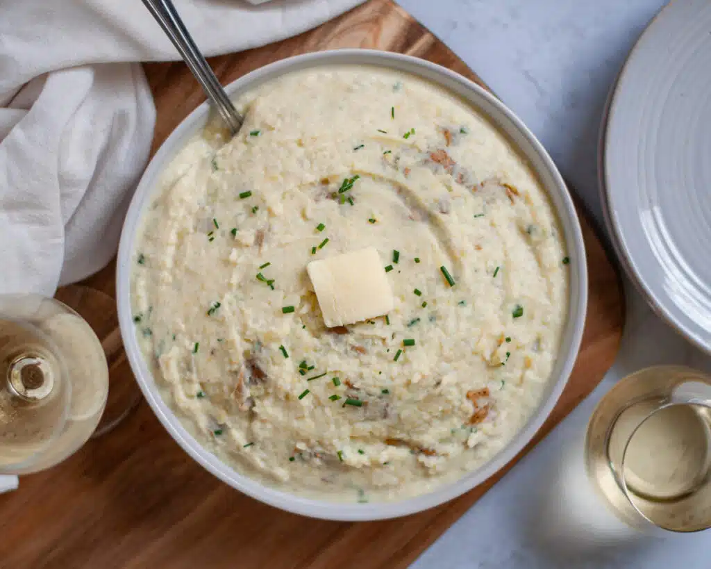 A bowl of creamy mashed cauliflower topped with a pat of butter and garnished with chopped chives. A spoon is placed inside the bowl, and a glass of white wine is nearby on a wooden surface. A white cloth napkin and plate are partially visible.