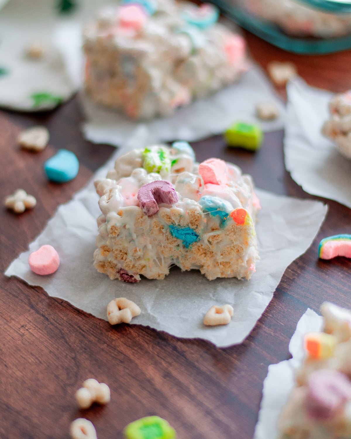 Lucky charm cereal bars set out on pieces of parchment paper and surrounded by additional cereal and colorful marshmallows.