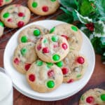 A plate full of Christmas m&m Christmas cookies, with more cookies, a glass of milk, and holly around the scene.
