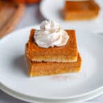 Close up of a stack of two pumpkin pie bars topped with a dollop of whipped cream on a white serving plate.