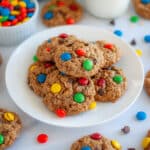 A plate stacked with monster cookies, with additional cookies, m&ms, chocolate chips, and oats around it.