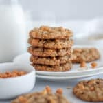 A stack of cookies on a white serving plate, with additional cookies, butterscotch chips, and oats around it.