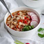A white serving bowl filled with red pozole soup and garnished with radishes and cilantro.