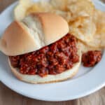 Close up of a sloppy joe served on a bun with potato chips on a white plate.