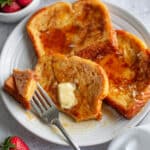 Close up of a plate of French toast smothered in butter and maple syrup, showcasing a fork holding a bite ready to eat.