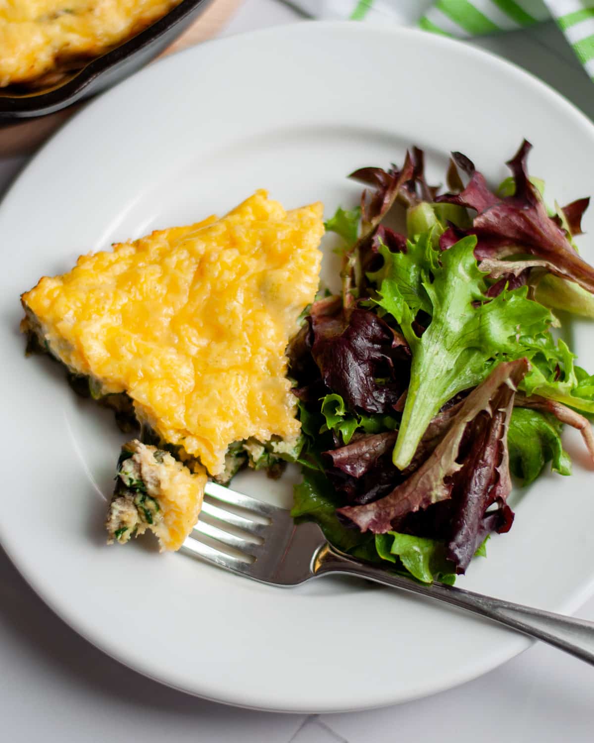 A slice of asparagus frittata, along with a fork with a bite removed, on a plate with a side salad.