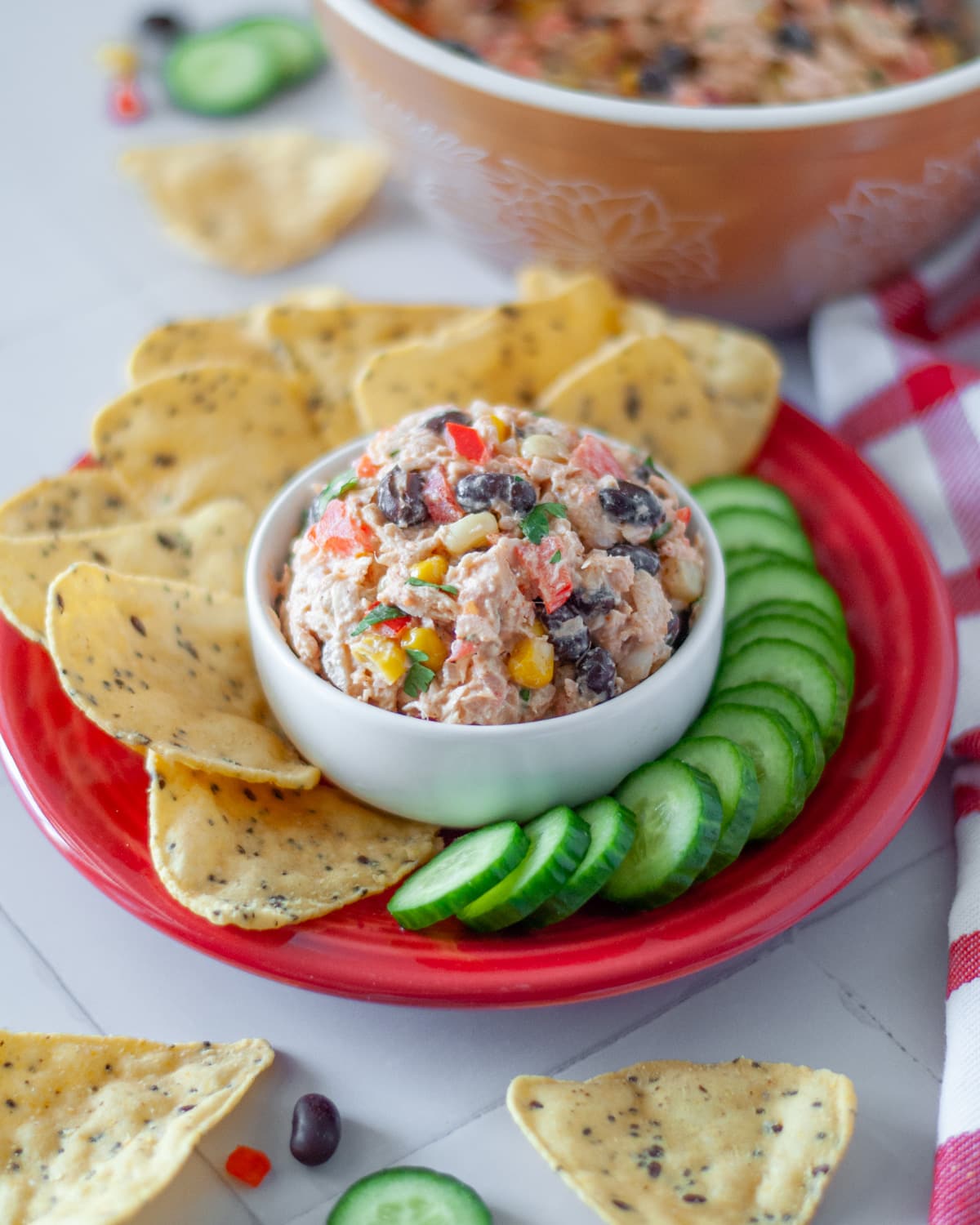A small bowl of Mexican tuna salad set on a plate with tortilla chips and cucumber slices.