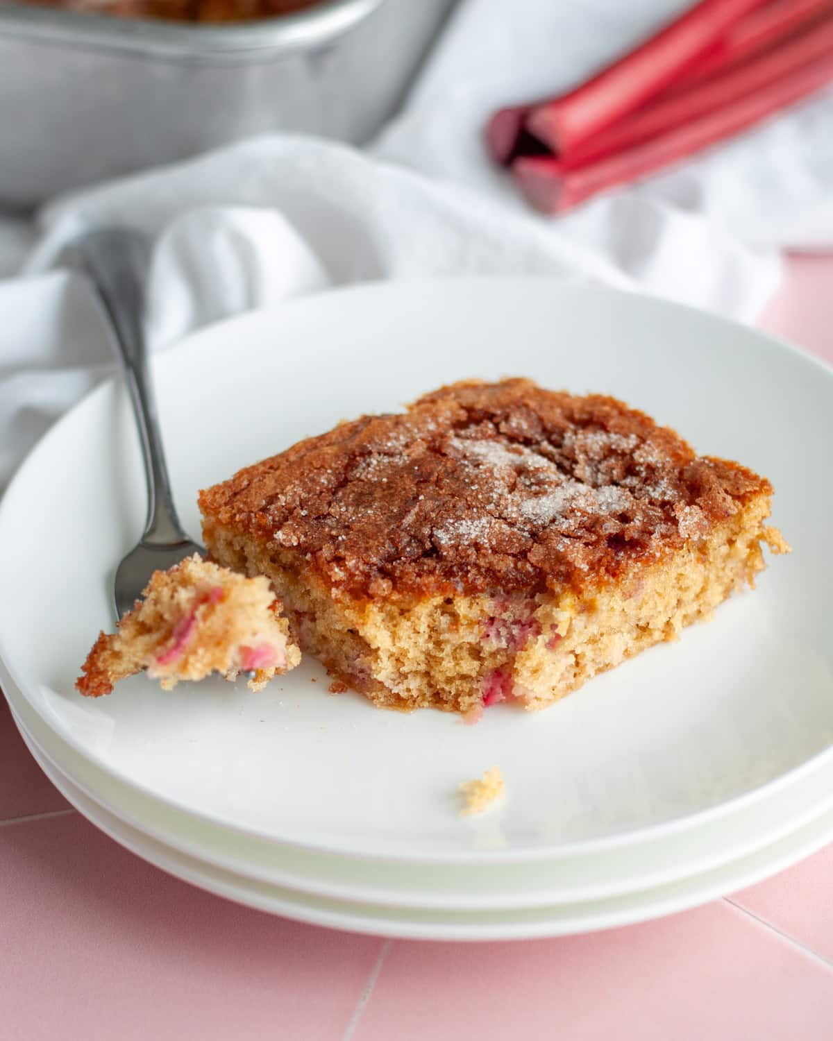 Close up of a slice of rhubarb cake with a bite taken out of it on a fork that's set on a stack of white plates.
