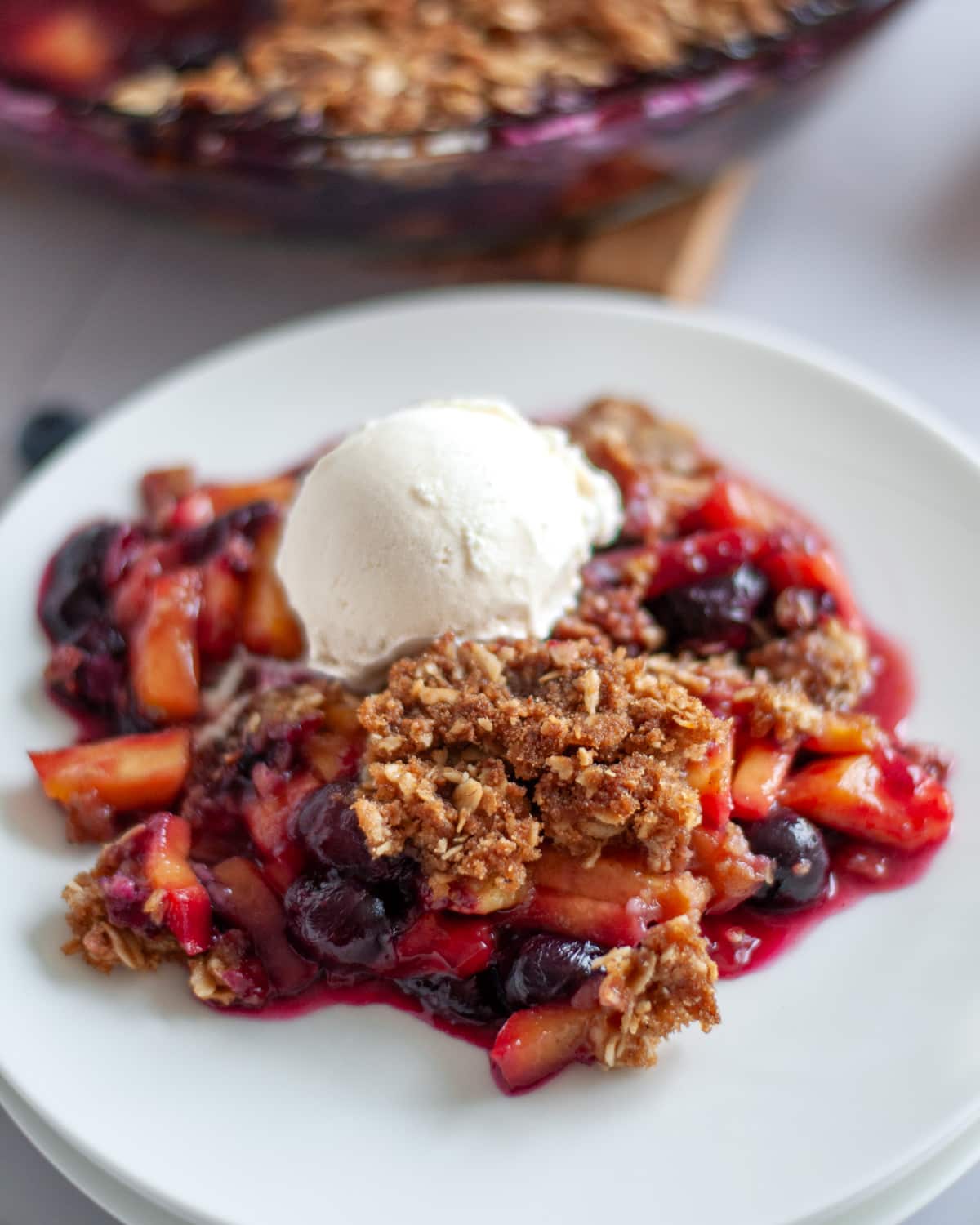 A scoop of peach and blueberry crisp topped with a scoop of vanilla ice cream on a white serving plate.