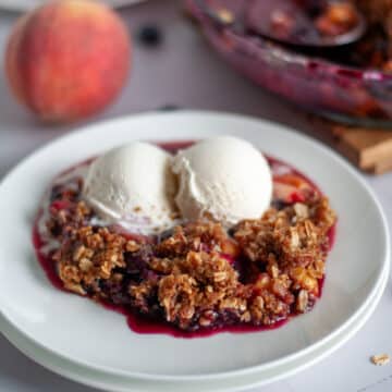 A scoop of peach and blueberry crisp topped with a couple of small scoops of vanilla ice cream on a white serving plate.
