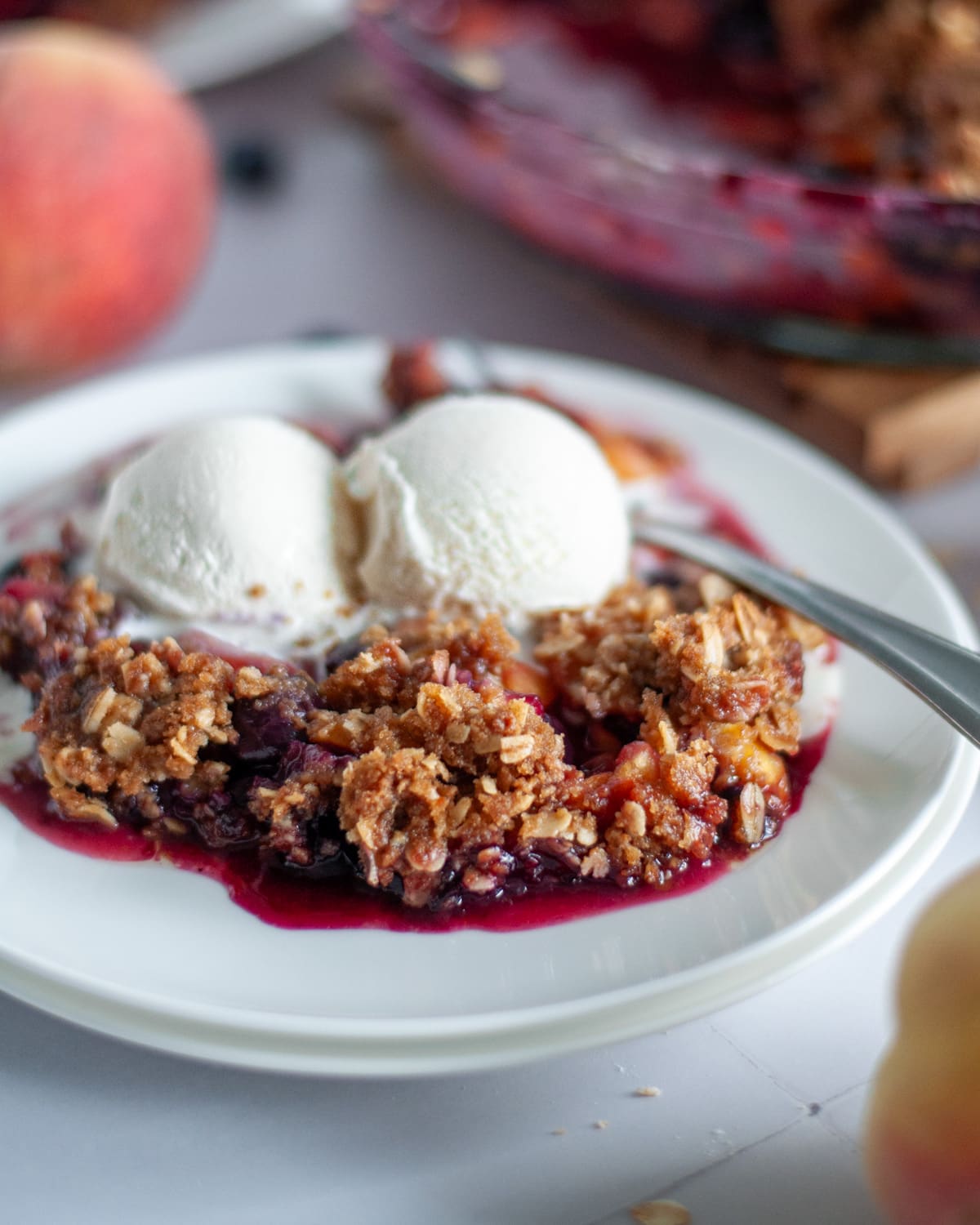 A scoop of peach and blueberry crisp topped with a couple of small scoops of vanilla ice cream on a white serving plate.