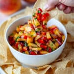 A hand with a tortilla chip dunked into the peach salsa, ready to eat it.