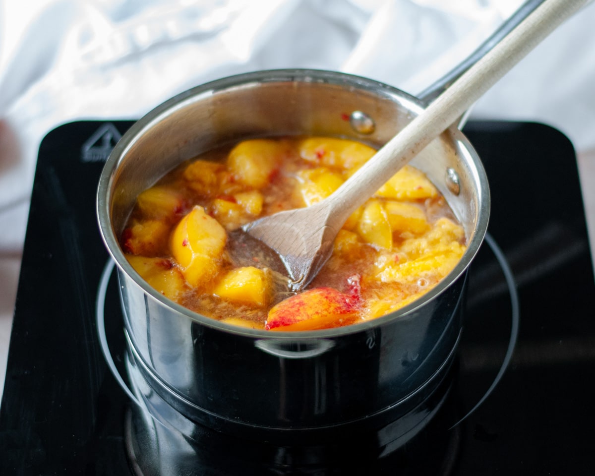 Process shot showing the peach syrup ingredients while cooking, with the peaches being broken down with a wooden spoon.