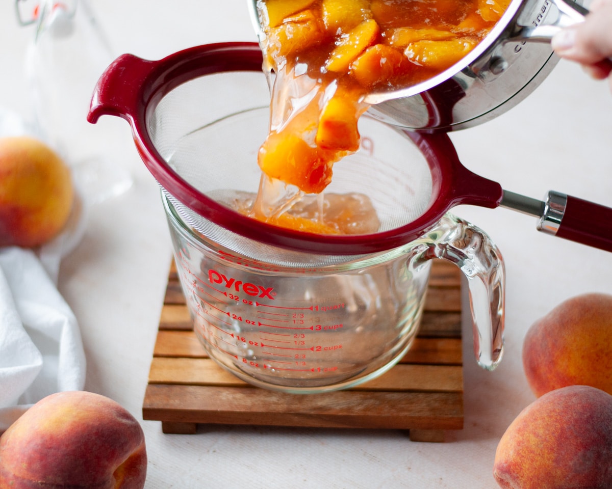 Process shot showing peach syrup being poured into a fine mesh strainer on a large liquid measuring cup to remove the fruit.