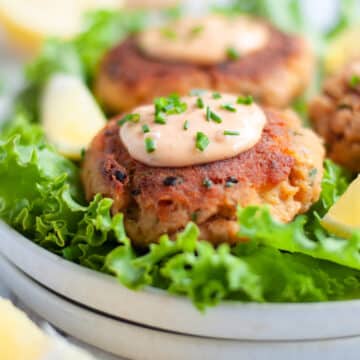 Close up of a plate of smoked salmon patties siting on lettuce leaves next to lemon wedges, topped with a sauce and fresh chives.